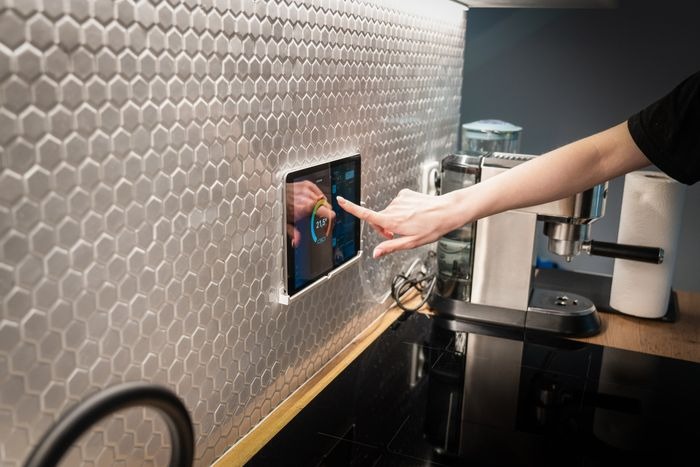 A person taps a wall-mounted smart home control panel in a modern kitchen, with a hexagon tile backsplash, espresso machine, and glossy black countertop visible.